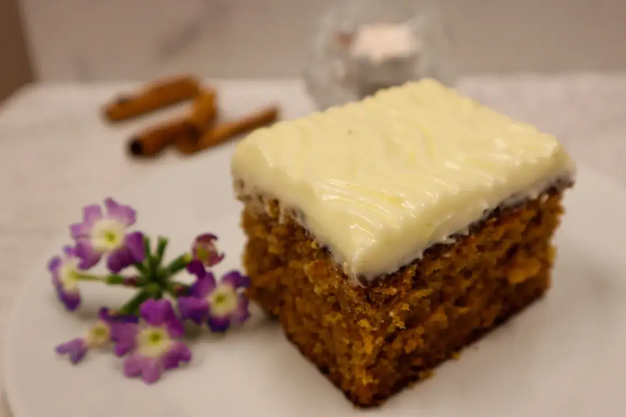 En bit saftig glutenfri morotskaka med krämig frosting, dekorerad med lila blommor och kanelstänger i bakgrunden.