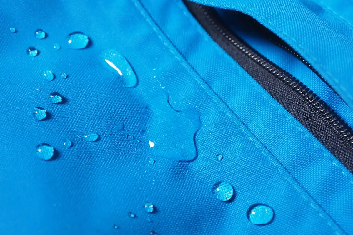 Gotas de agua sobre tela azul de poliamida que abriga, mostrando su resistencia al agua. Cremallera negra visible.