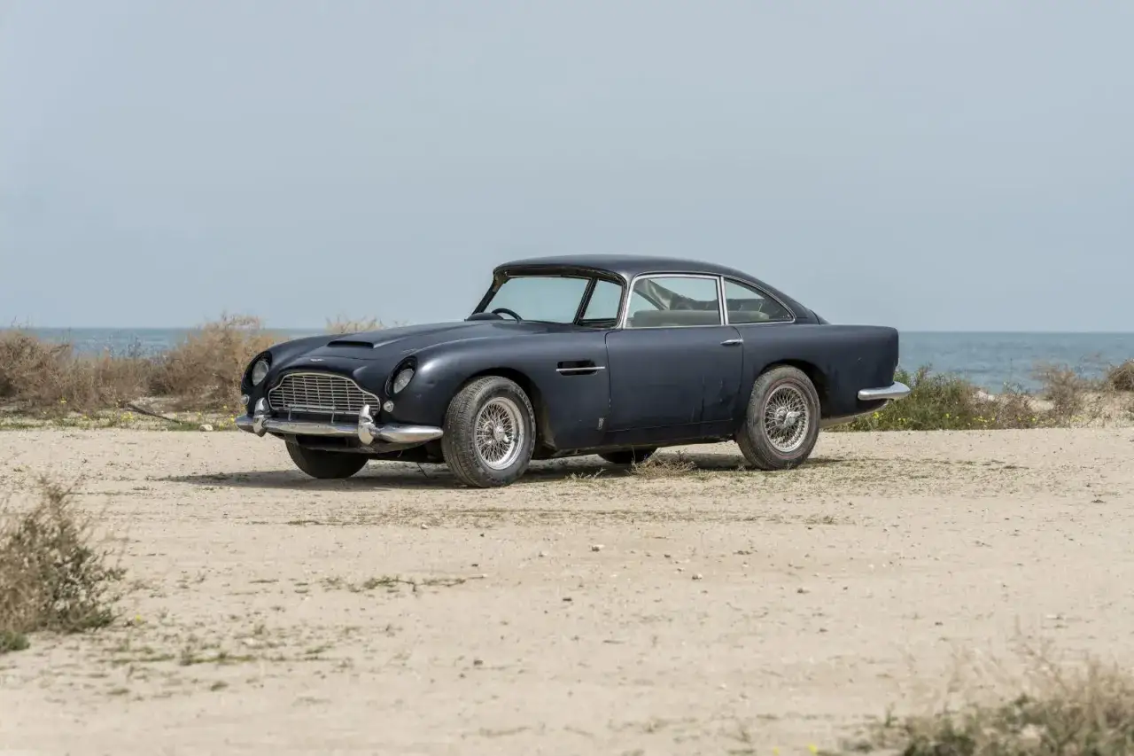 Klasyczny Aston Martin DB5 na piaszczystym pustkowiu, z morzem w tle.