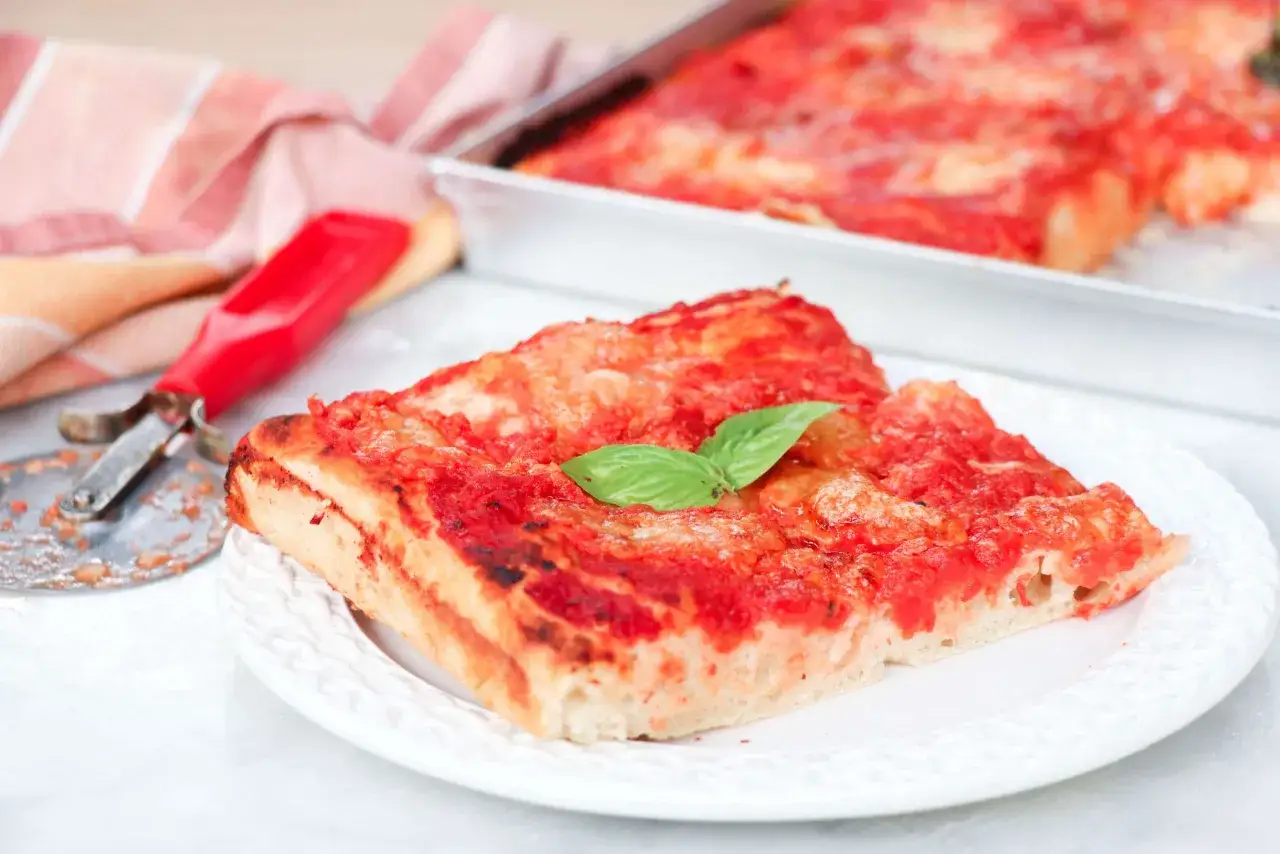 Una fetta di pizza fatta in casa teglia, con pomodoro e basilico, accanto a un tagliapizza rosso.