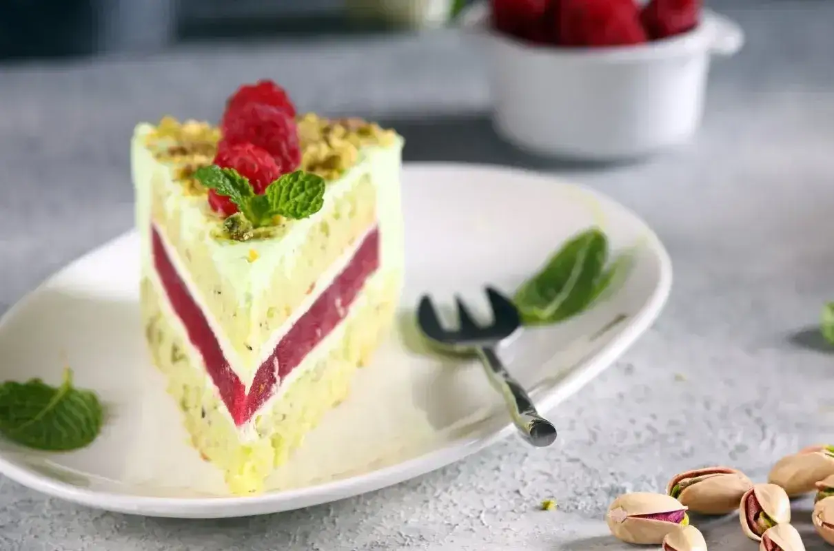 Pyszny tort pistacjowo malinowy z malinami i miętą na talerzu. Obok widoczne łuskane pistacje.