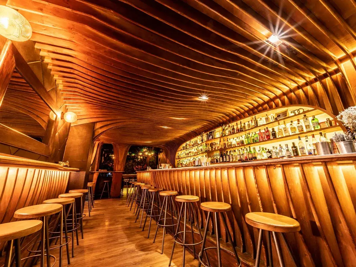 Interior de un gastro bar con techo y paredes de madera ondulada, barra con taburetes y estanter&iacute;as repletas de botellas.