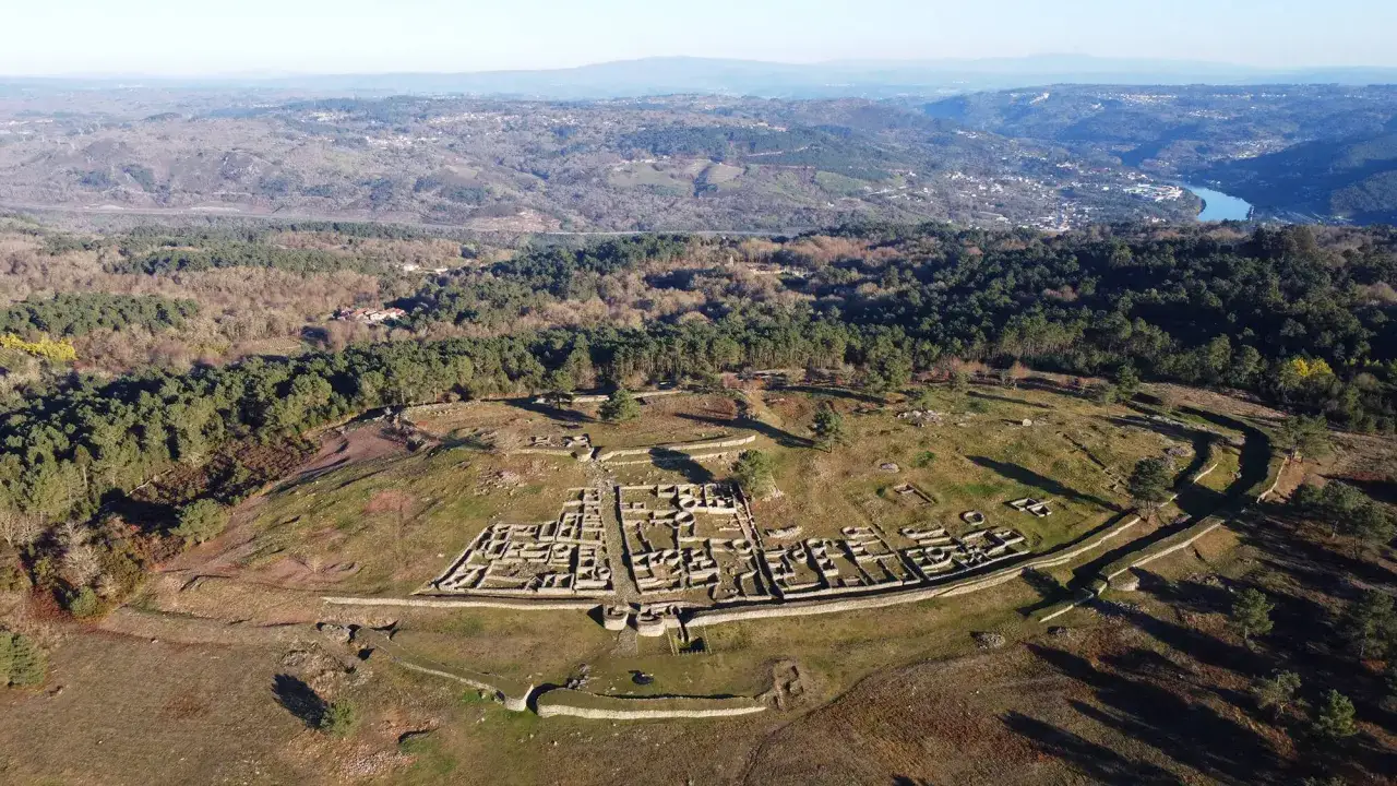 Vista aérea del castro de Santa Tegra, con sus murallas y edificaciones de piedra, rodeado de bosques y con un valle al fondo.