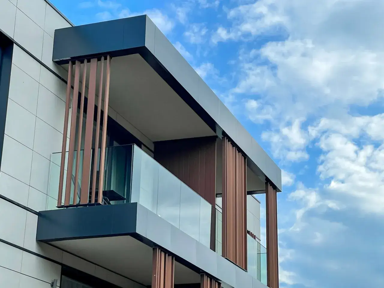 Balc&oacute;n moderno con barandilla de vidrio laminar de seguridad y listones de madera, bajo un cielo azul con nubes.