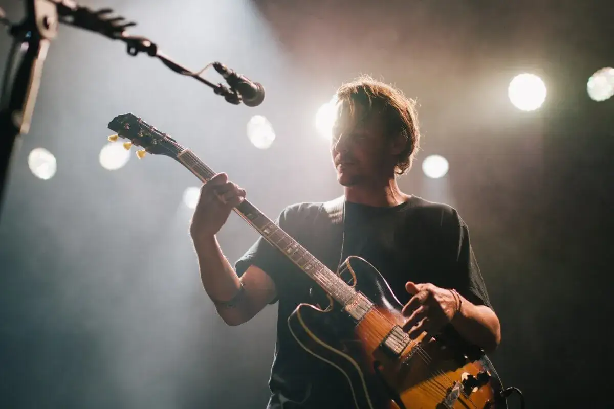 Mężczyzna gra na gitarze elektrycznej, jeden z wykonawców muzyki alternatywnej, na tle scenicznych świateł.