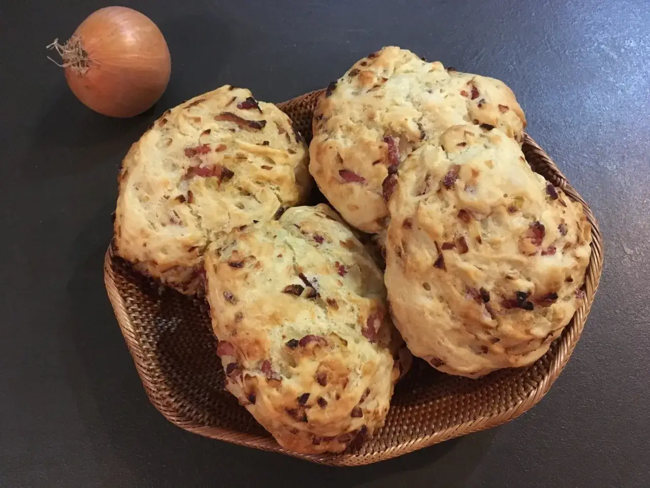 Un panier tress&eacute; contient quatre pains aux lardons et oignons dor&eacute;s. Un oignon cru repose &agrave; c&ocirc;t&eacute;.