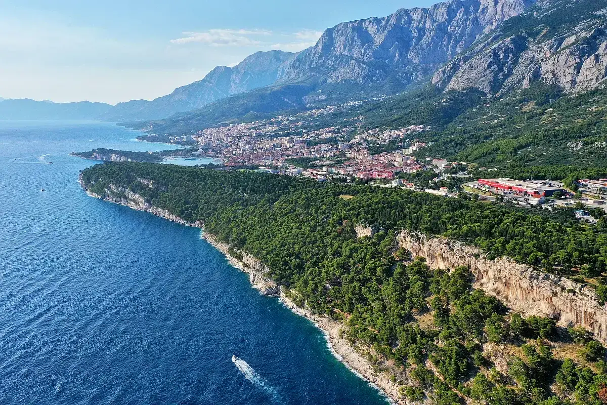 En vy över Makarska strandpromenad med turkost hav, gröna pinjeskogar och berg i bakgrunden. En båt syns på vattnet.
