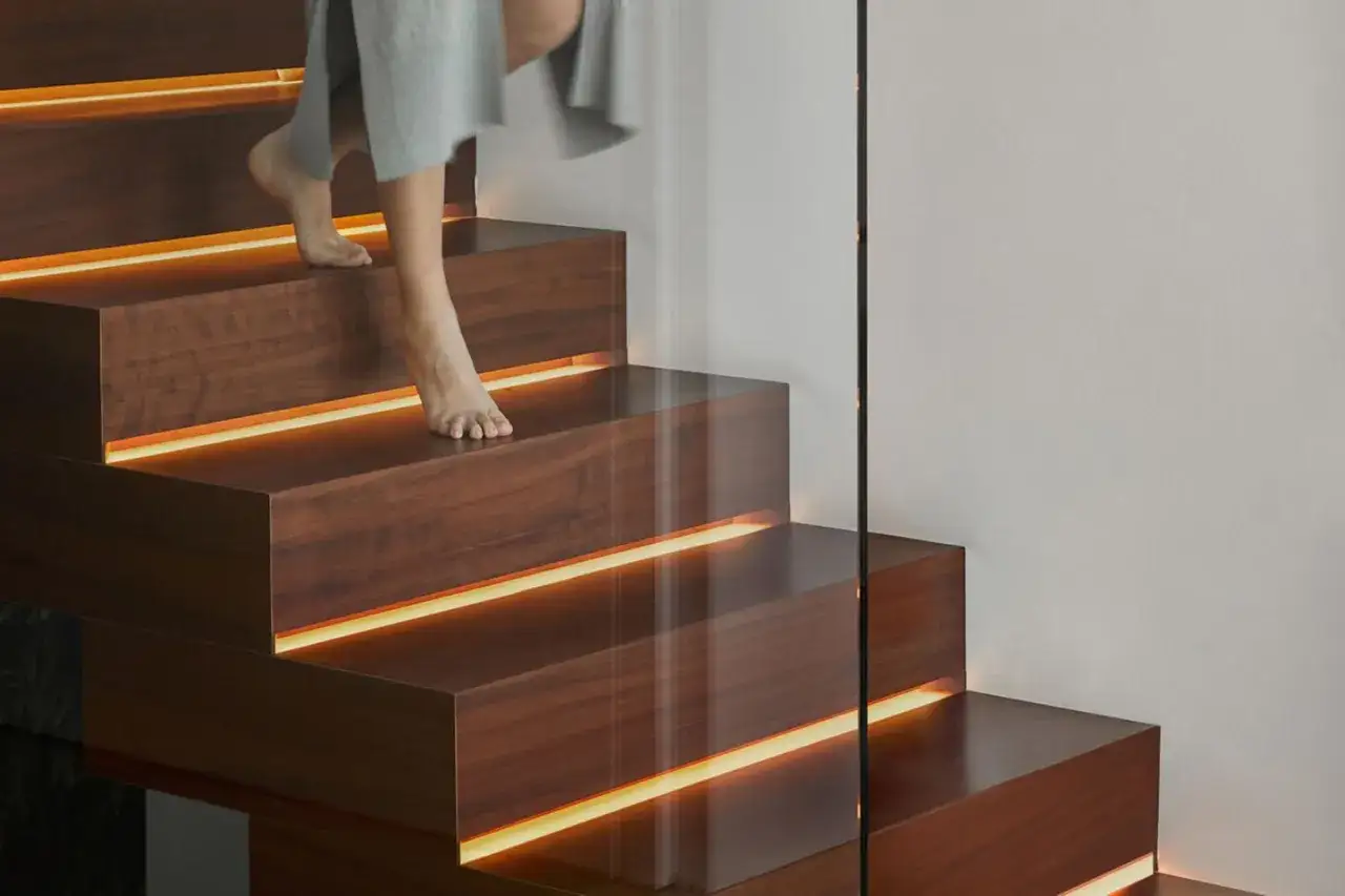 Ein modernes Treppenhaus gestalten: Holzstufen mit indirekter Beleuchtung und Glasgeländer. Barfuß geht es die beleuchtete Treppe hinauf.