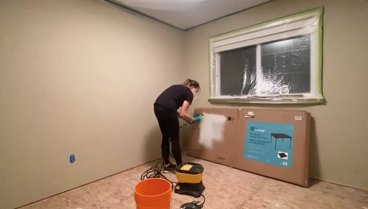 Donna imbianca a spruzzo una parete, con un secchio arancione e una macchina per verniciatura a spruzzo sul pavimento.