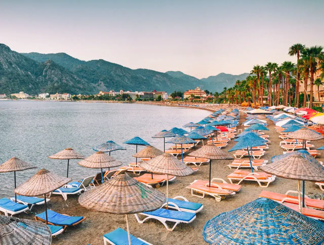 Plaża w Marmaris, Turcja, z rzędami leżak&oacute;w i parasoli, w tle g&oacute;ry i hotele.