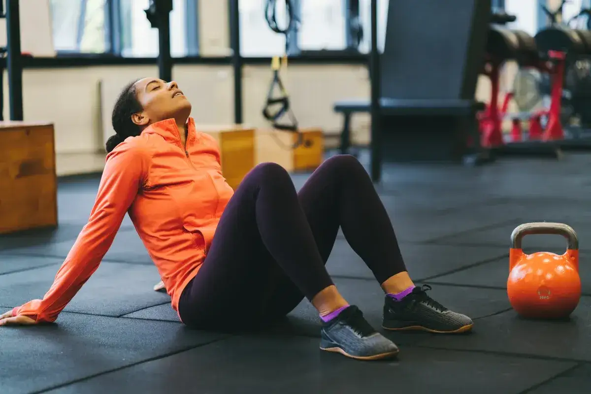 Frau sitzt erschöpft auf dem Boden im Fitnessstudio, neben einer Kettlebell. Sie ist auf dem Weg, um abzunehmen in 4 Wochen.