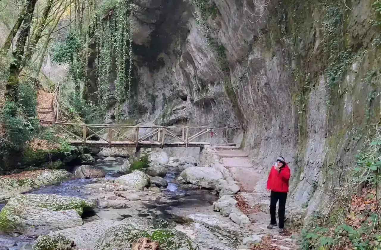 Una persona in giacca rossa scatta una foto nella **valle dell'Orfento**, con un ponte di legno e un ruscello.