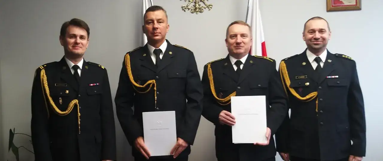Czterech strażaków w galowych mundurach odbiera dokumenty, symbolizujące ich emeryturę.