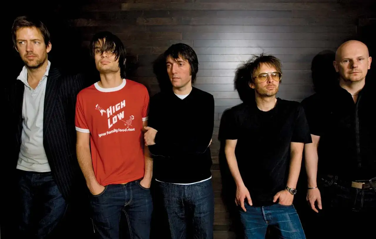 Die Mitglieder von Radiohead stehen nebeneinander vor einer dunklen Holzwand. Einer tr&auml;gt ein rotes T-Shirt mit der Aufschrift 
