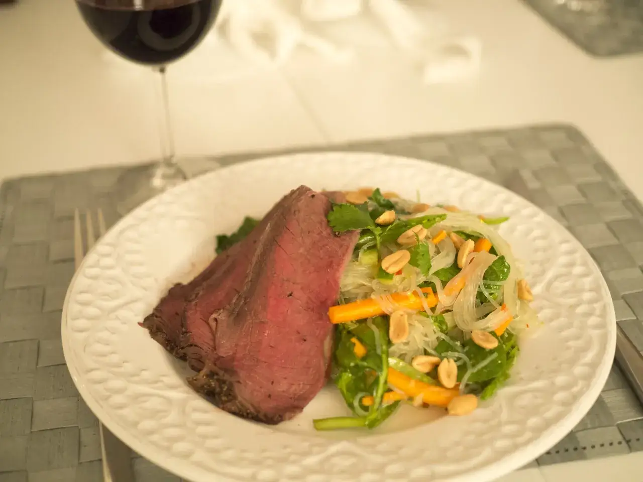 En saftig stek serveras med en fr&auml;sch asiatisk biffsallad med glasnudlar, gr&ouml;nsaker och jordn&ouml;tter. Ett glas r&ouml;dvin st&aring;r bredvid.