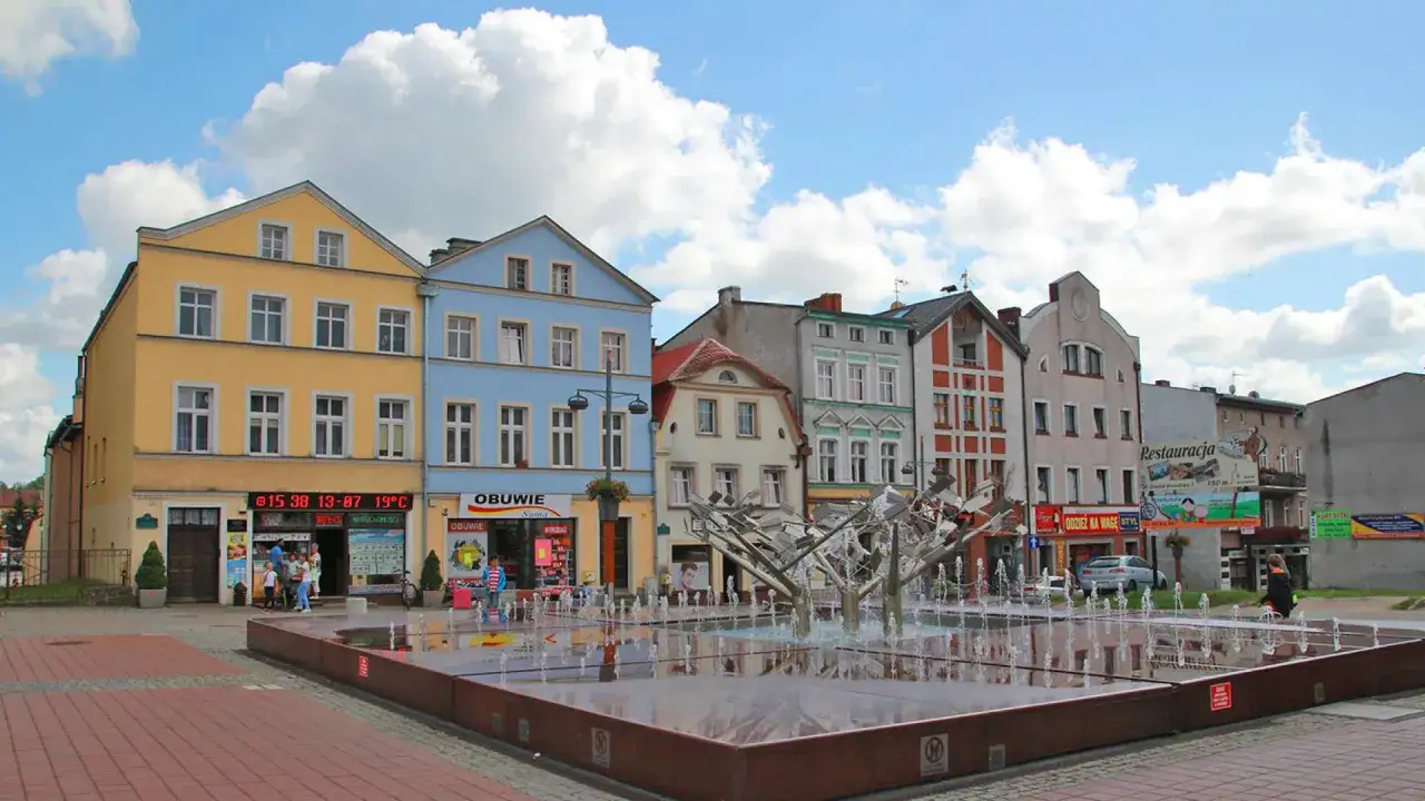 Kolorowy rynek w Bytowie z fontanną i kolorowymi kamienicami. W tle widoczne sklepy i restauracja.
