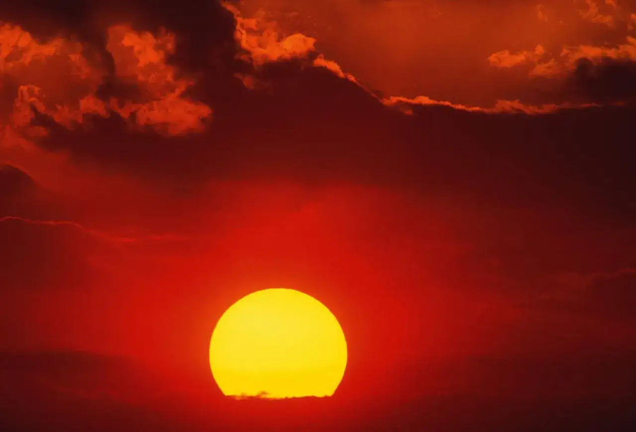 Le soleil, une boule jaune éclatante, se couche dans un ciel rouge intense, illuminant les nuages d'une lueur spectaculaire.