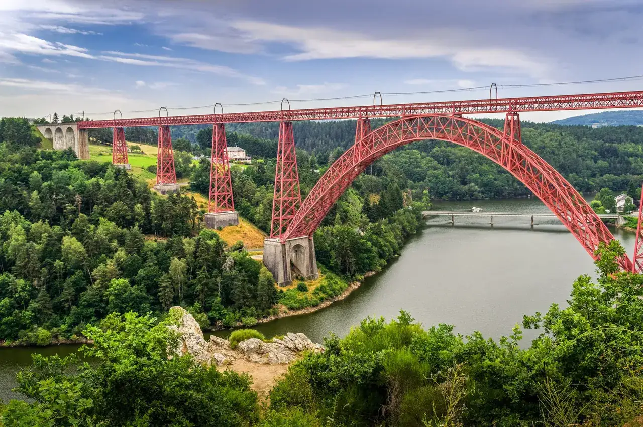 Impressionnant viaduc rouge traversant les gorges de la Truyère, un paysage verdoyant et un lac paisible.