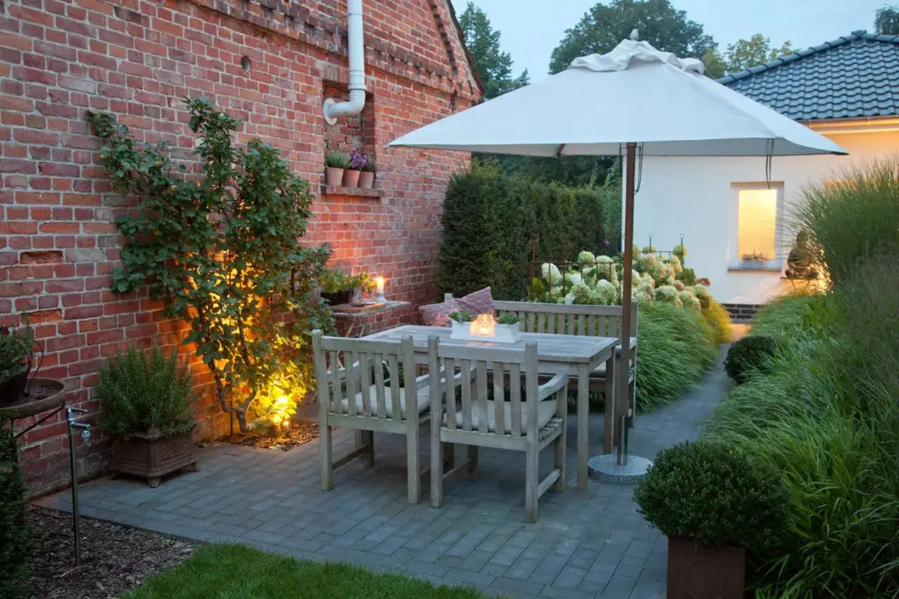 Gem&uuml;tliche Holzm&ouml;bel auf gepflasterter Fl&auml;che laden ein, die Terrasse gestalten. Ein Sonnenschirm spendet Schatten, w&auml;hrend Pflanzen und Beleuchtung f&uuml;r Ambiente sorgen.