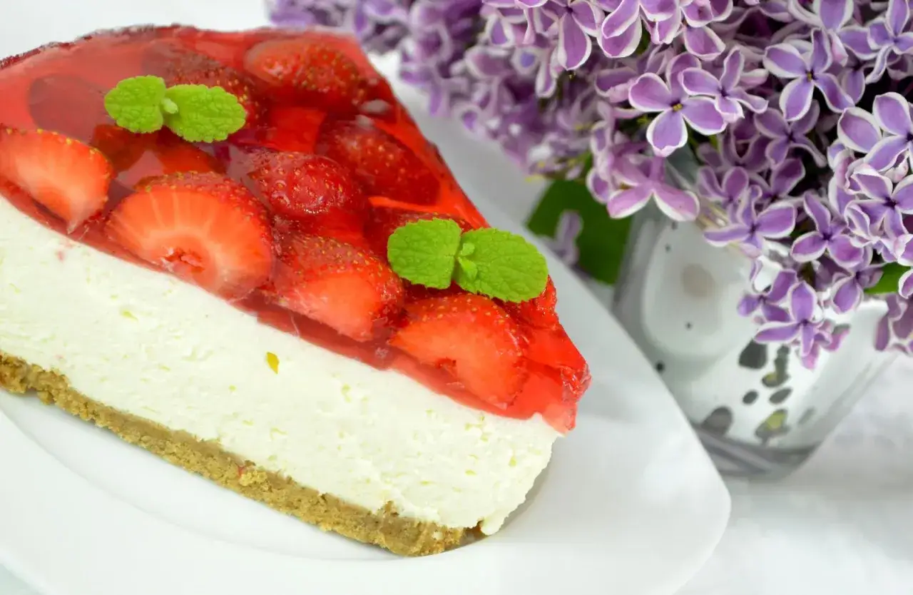 Pyszny sernik na zimno z mascarpone, udekorowany truskawkami i galaretką, obok bukiet bzu.