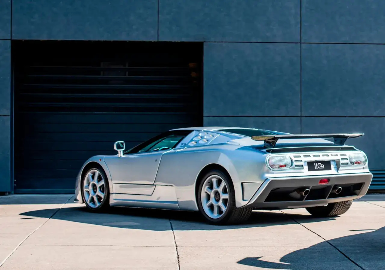 Srebrny Bugatti EB110 z charakterystycznym spoilerem i wlotami powietrza, zaparkowany przed nowoczesnym budynkiem.