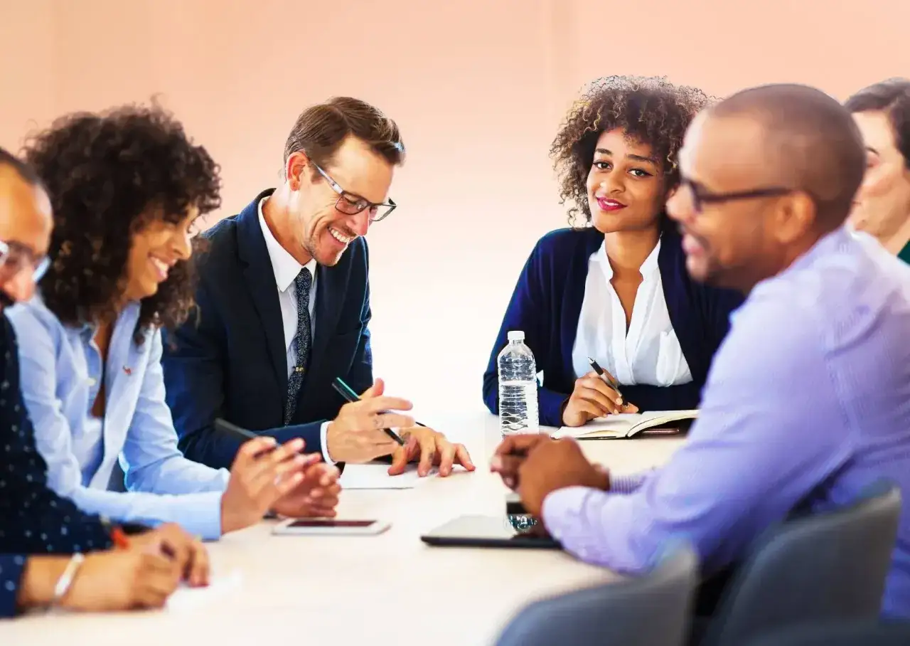 A diverse leader smiles during a meeting, surrounded by colleagues engaged in discussion.