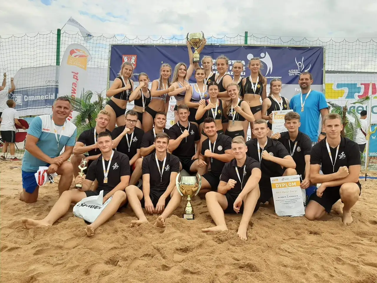Drużyna MKS Mazur Sierpc świętuje zwycięstwo na plaży, trzymając puchary i dyplomy.