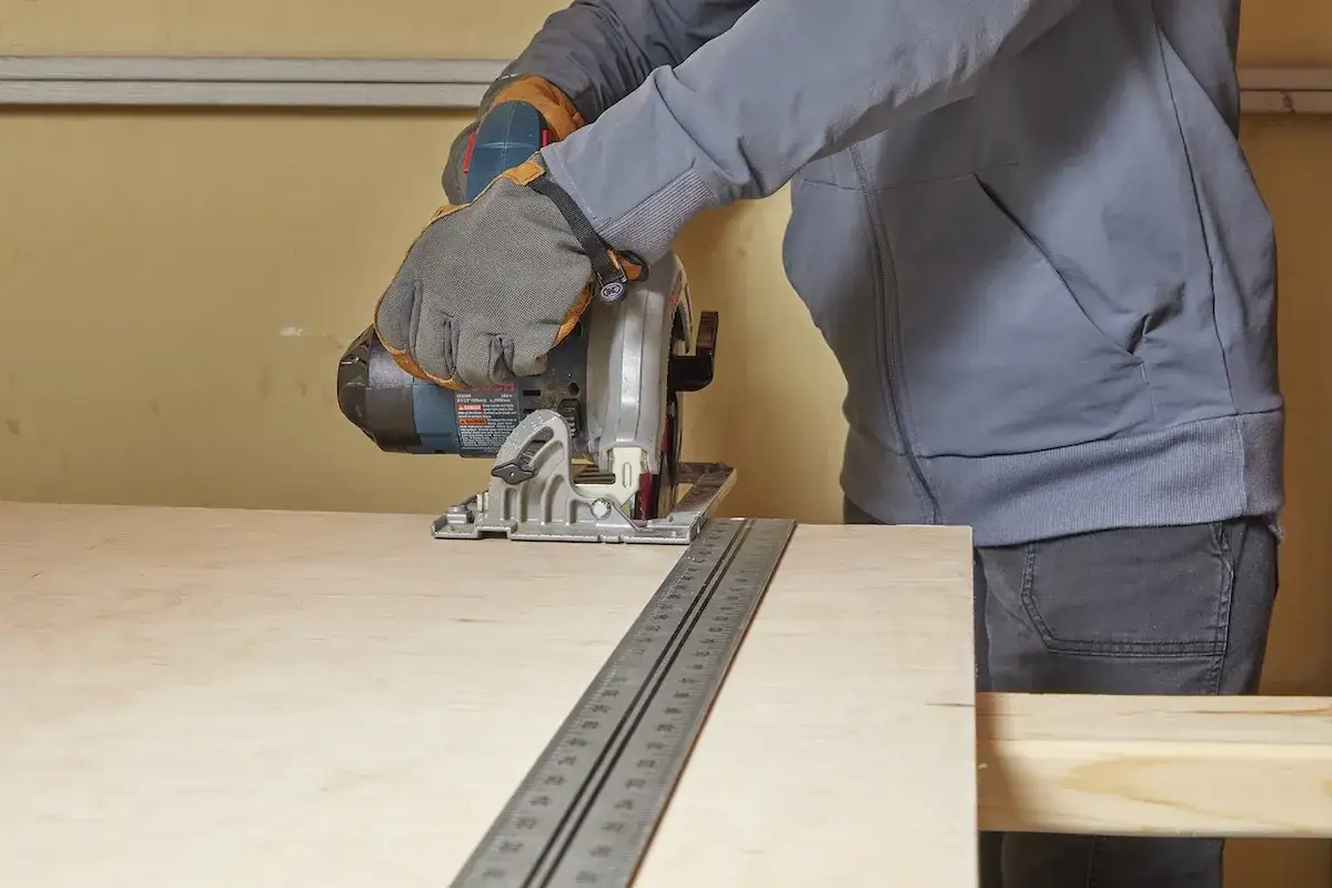 Persona con guanti sta usando una sega circolare per tagliare compensato, guidata da un righello metallico per una linea precisa.