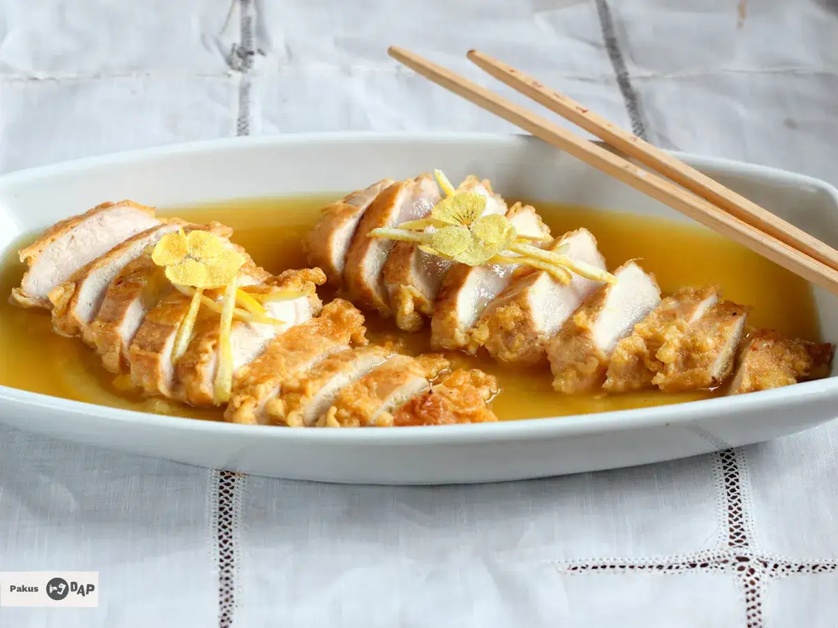 Delicioso pollo al limón, crujiente por fuera y tierno por dentro, bañado en una salsa dorada y decorado con finas hebras de limón y una flor amarilla.