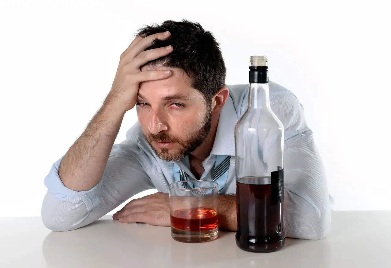 Zrezygnowany mężczyzna z ręką na czole, obok butelka i szklanka z alkoholem. Czy to już 13.7 promila?