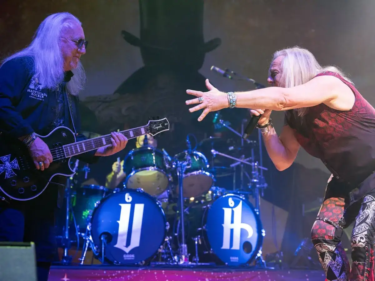 Uriah Heep członkowie na scenie: gitarzysta z długimi białymi włosami gra na czarnej gitarze, wokalista śpiewa do mikrofonu.