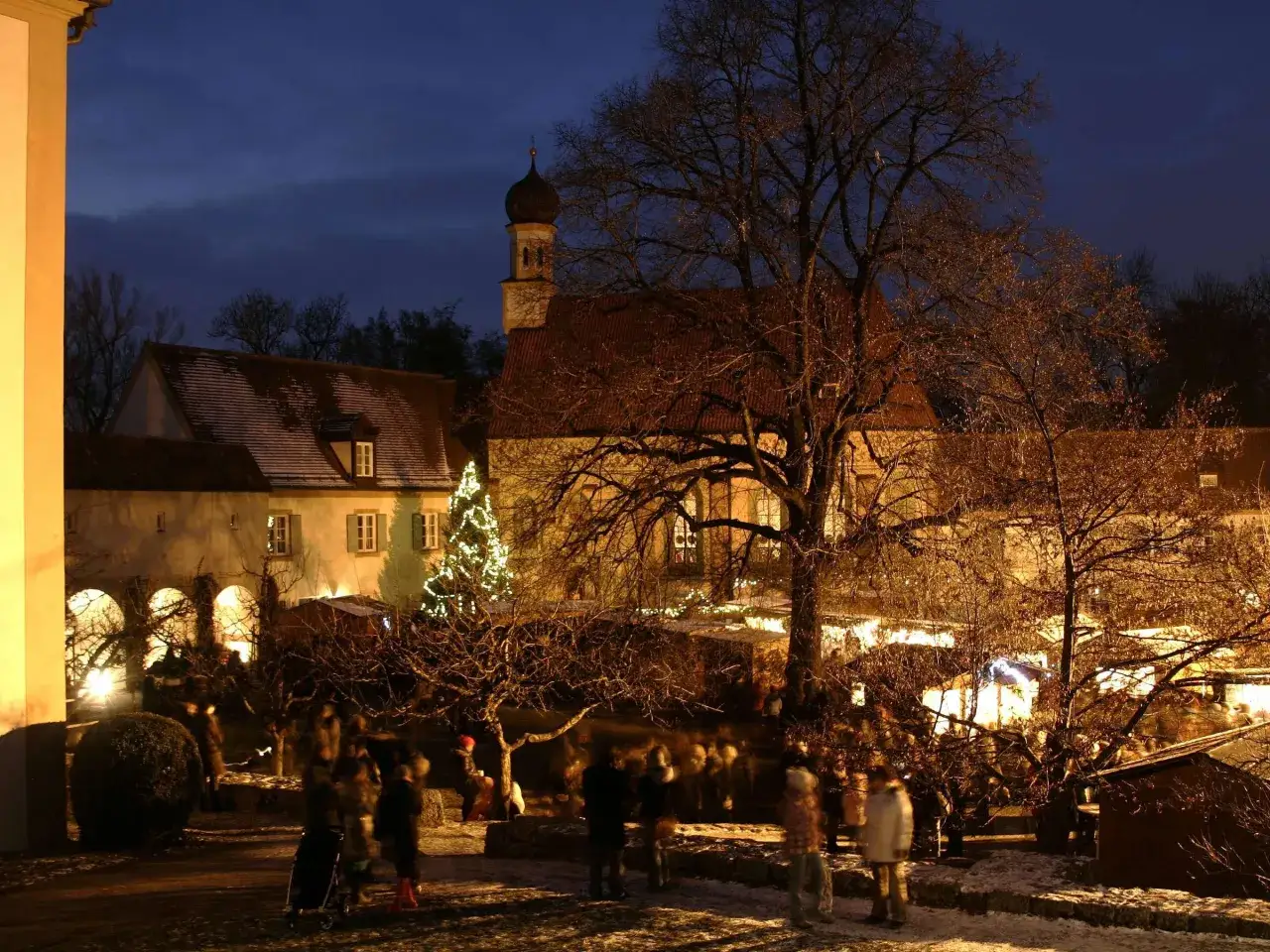 Besucher genie&szlig;en den stimmungsvollen schloss nymphenburg weihnachtsmarkt im Abendlicht, mit beleuchtetem Baum und historischen Geb&auml;uden.