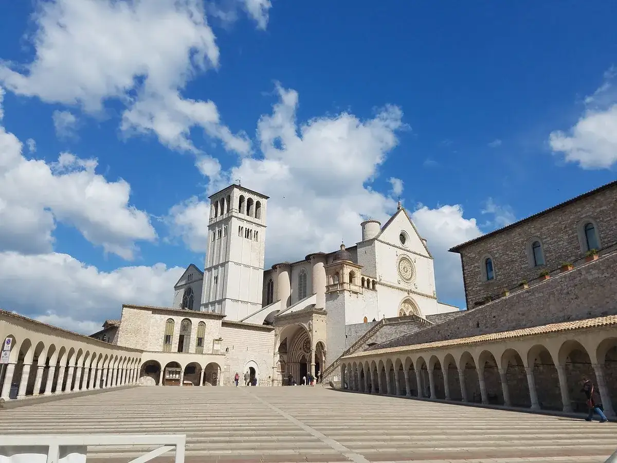 Piazza del **centro storico** di Assisi con la Basilica di San Francesco e il suo campanile sotto un cielo azzurro con nuvole bianche.