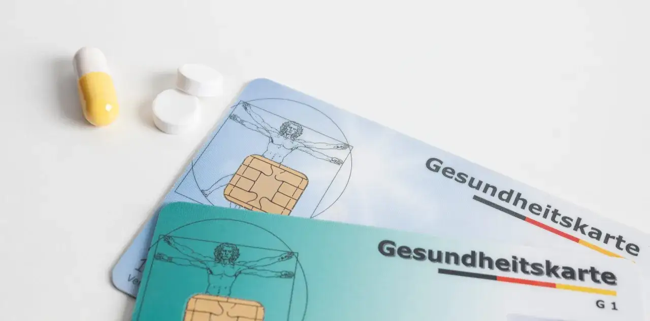 Zwei Gesundheitskarten, eine gelbe Kapsel und zwei weiße Tabletten liegen auf weißem Grund. Die Karten zeigen den Vitruvianischen Menschen und sind mit