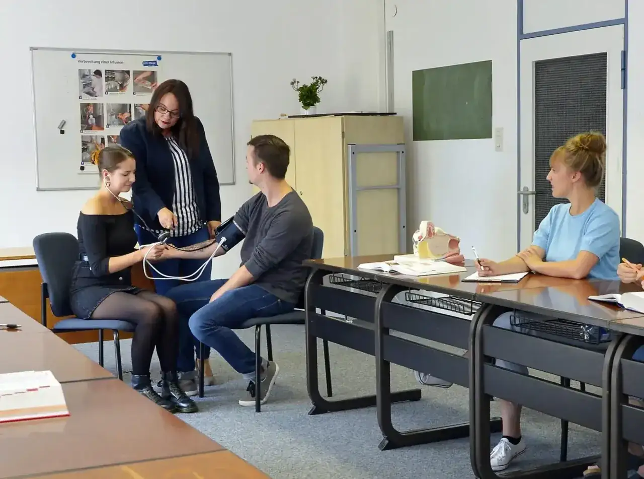 Sch&uuml;ler &uuml;ben Blutdruckmessen im Rahmen von **ausbildungen die nur 1 jahr dauern**. Eine Lehrerin erkl&auml;rt, w&auml;hrend eine Sch&uuml;lerin misst.