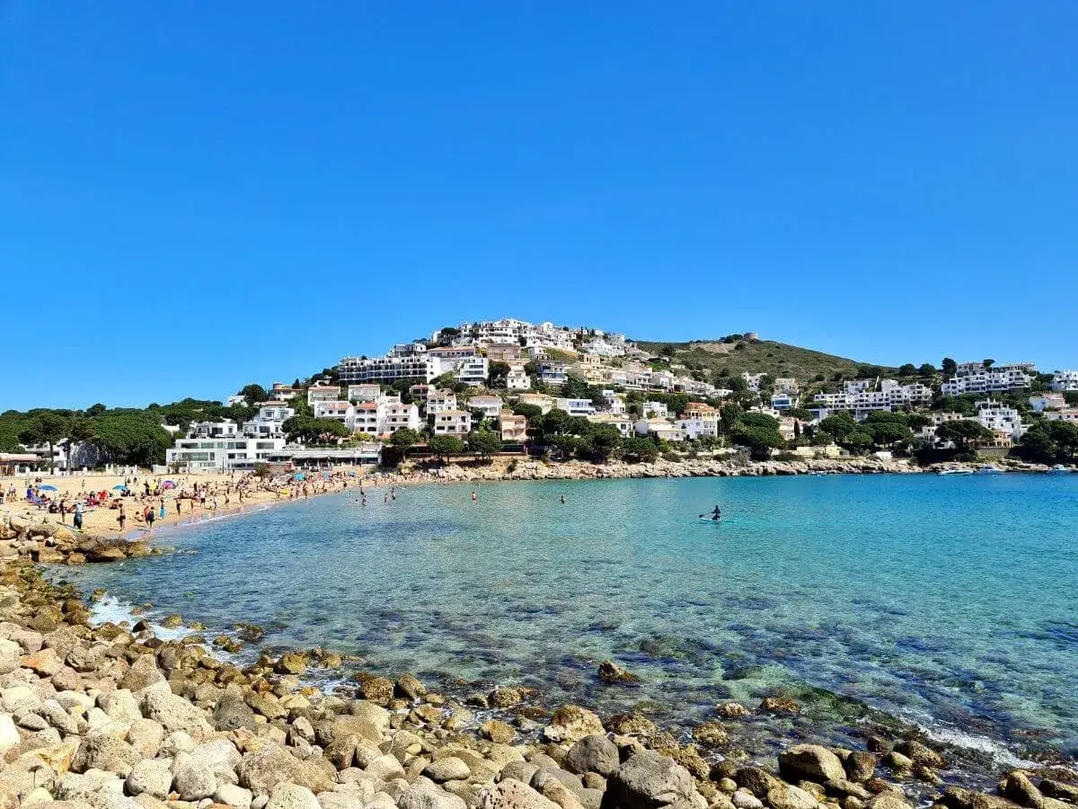 Vista de la platja de Montg&oacute;, con gente disfrutando del sol y el mar turquesa, y casas blancas escalonadas en la colina.