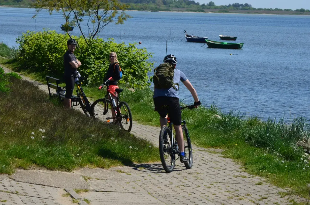 Trasa rowerowa Gdańsk Hel. Trzy osoby na rowerach odpoczywają nad wodą, podziwiając łodzie i piękny krajobraz.