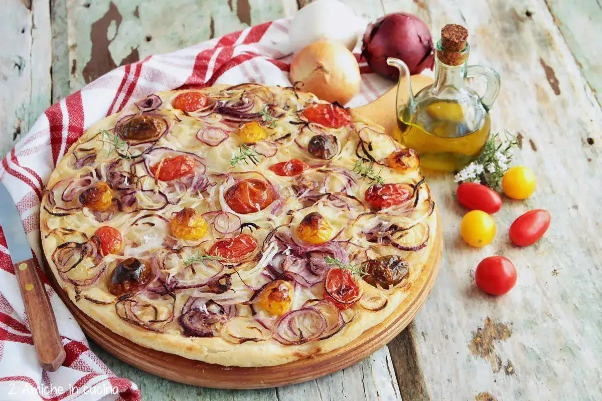 Focaccia con cipolle rosse e pomodorini, condita con olio d'oliva e erbe aromatiche, su un tavolo di legno rustico.