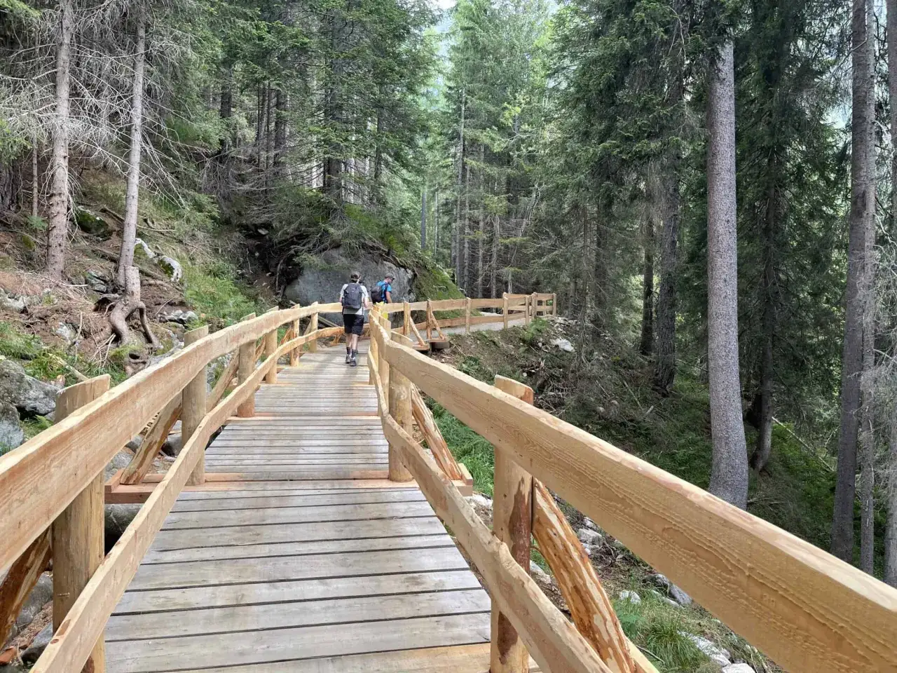 Sentiero in legno che conduce al lago Nambino, immerso in una foresta di conifere. Due escursionisti con zaini camminano sulla passerella.