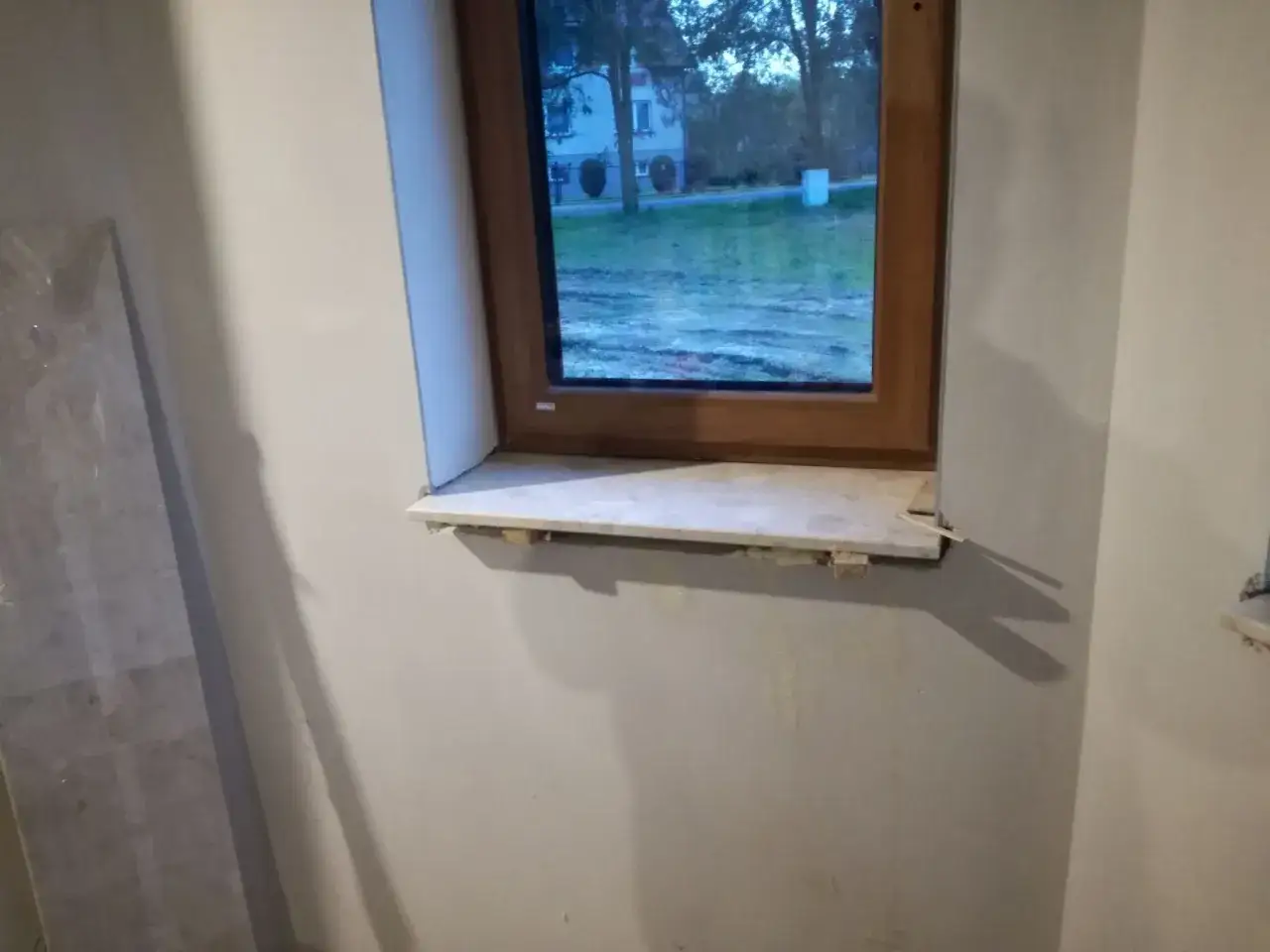 Początek montażu parapetu pod oknem. Widać drewnianą ramę okna i kamienny parapet podparty drewnianymi klockami.
