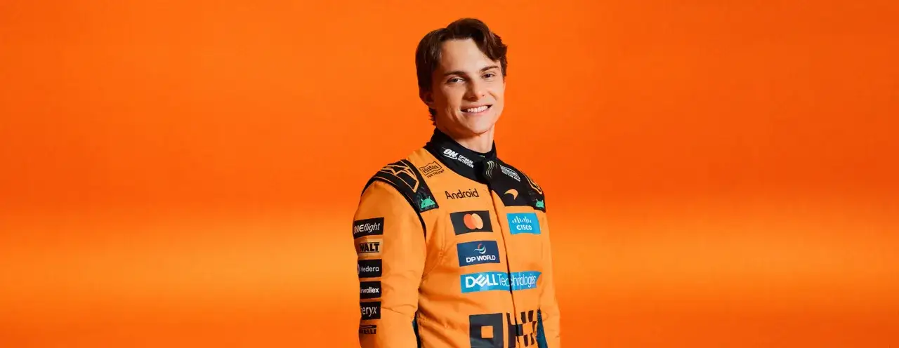 Joven piloto australiano de F1 con traje naranja de McLaren, sonriendo ante fondo naranja.