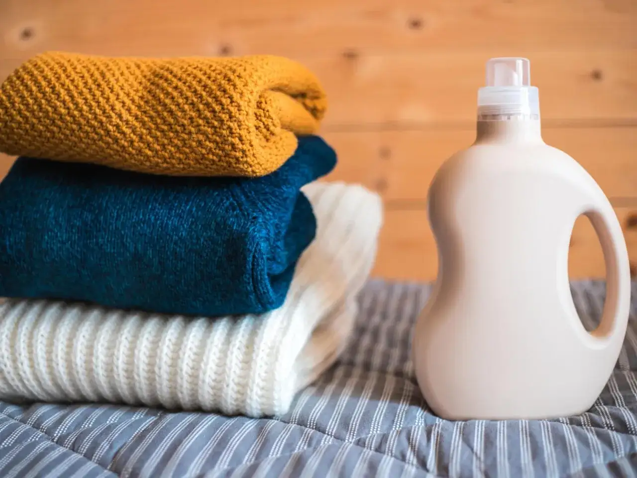 Ein Stapel gestrickter Pullover in Gelb, Blau und Weiß liegt neben einer Flasche Waschmittel. Perfekt für die **Wolle waschen**.