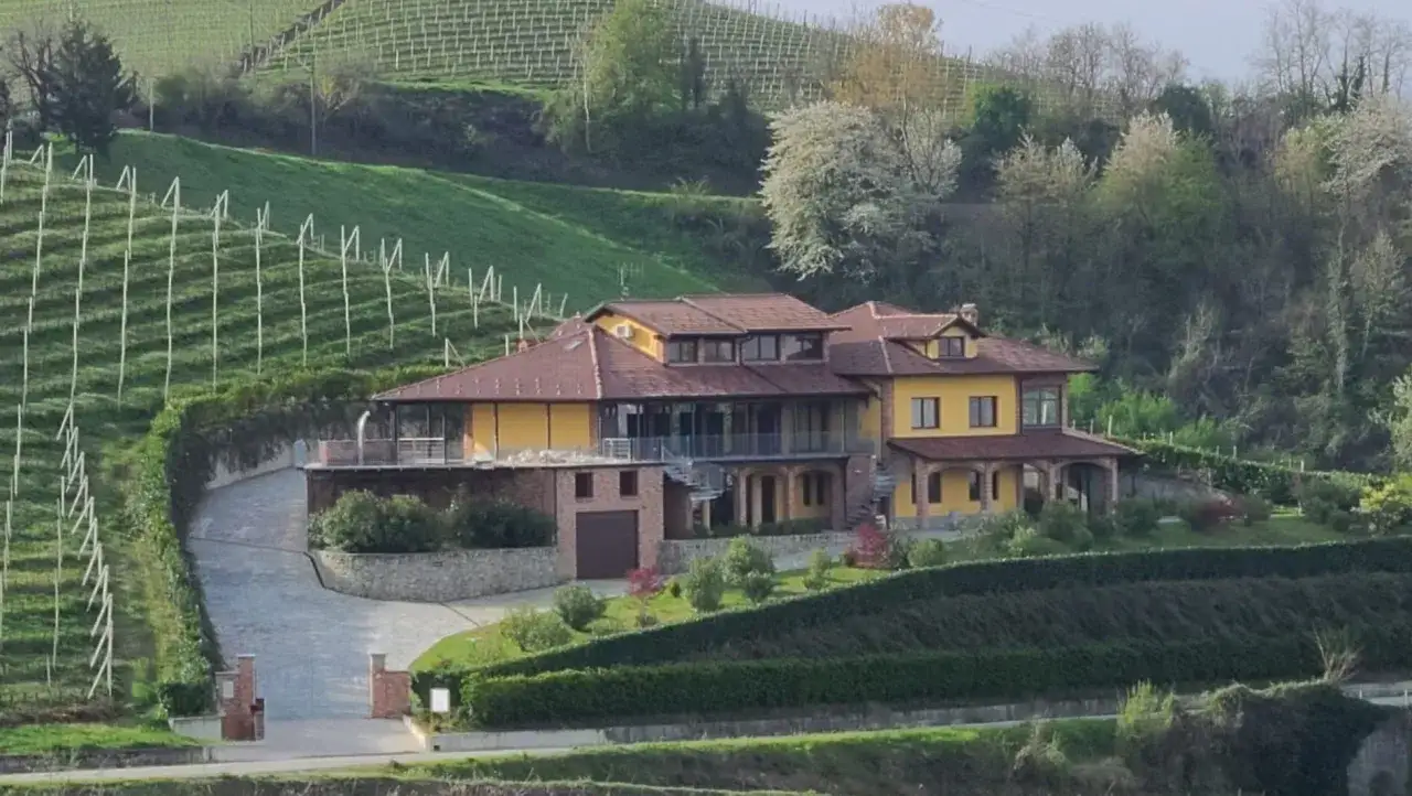 Villa gialla con tetto in tegole rosse, circondata da vigneti e alberi in fiore. Un vialetto conduce all'ingresso, ricordando un antico bricco maiolica.