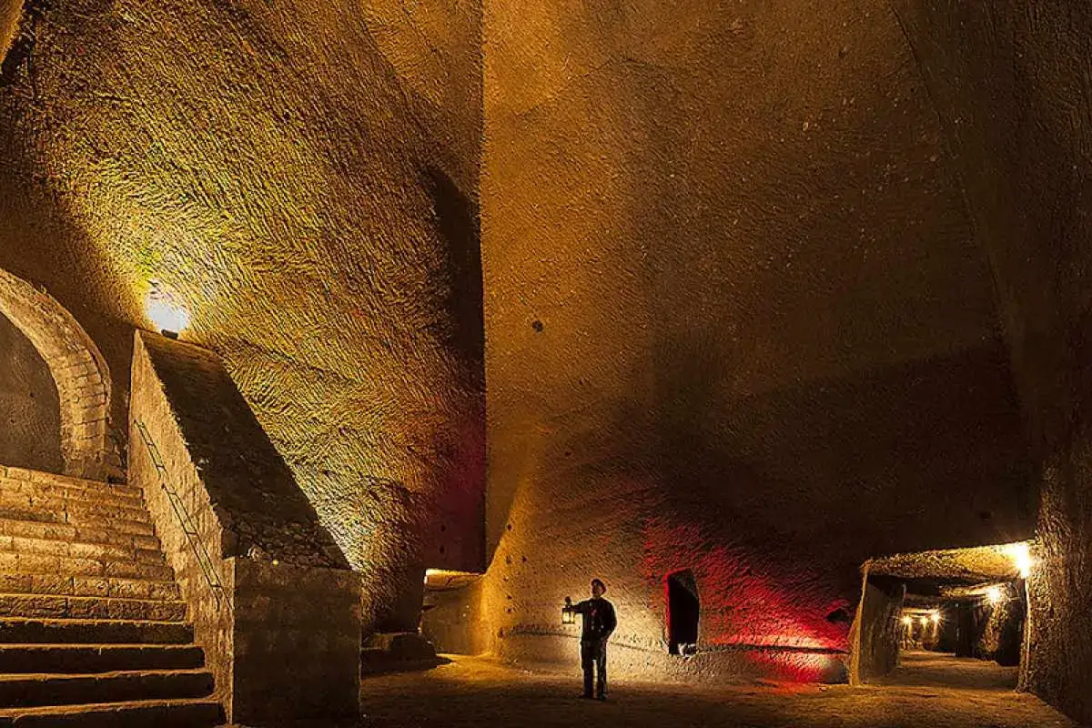 Un uomo con una lanterna esplora le suggestive cavità del Napoli Sotterranea, illuminato da luci dorate e rosse.