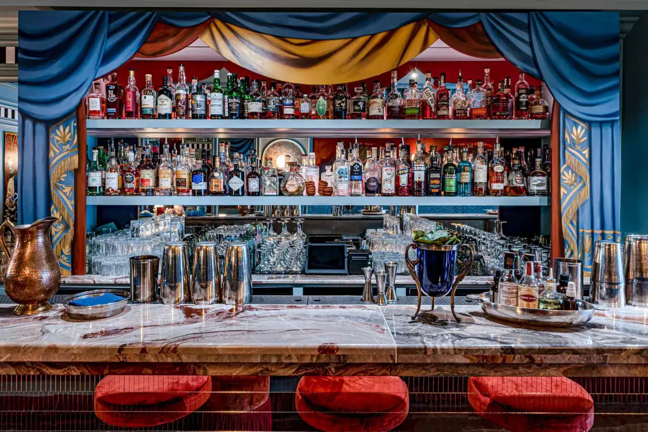 Un bar élégamment décoré avec des étagères remplies de bouteilles d'alcool variées. Des rideaux bleus et dorés encadrent la scène, invitant à un bisou bar.