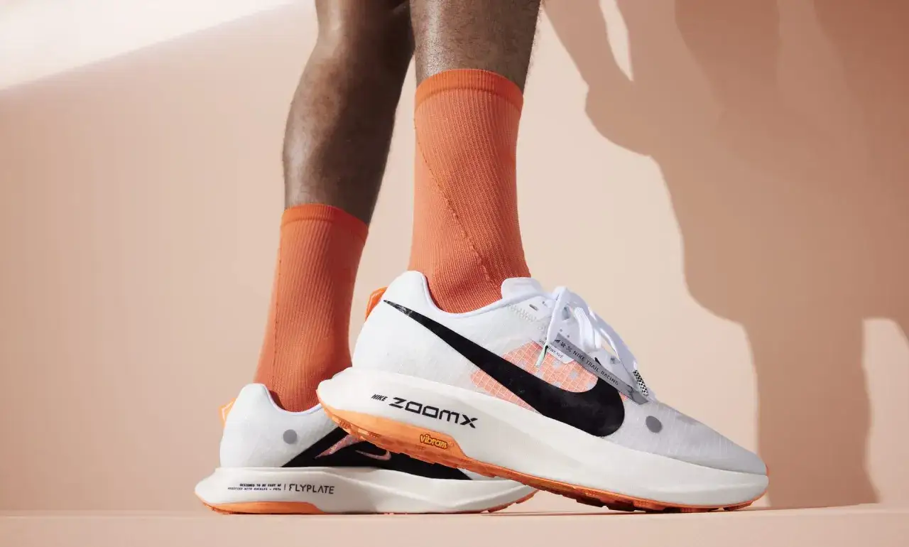 Białe buty Nike ZoomX z pomarańczowymi akcentami i czarnym logo na tle. Idealne do biegania.