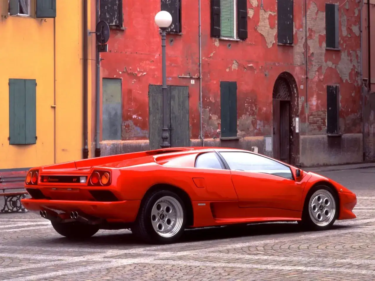 Czerwony Lamborghini Diablo stoi na brukowanym placu przed starym, czerwoną kamienicą.