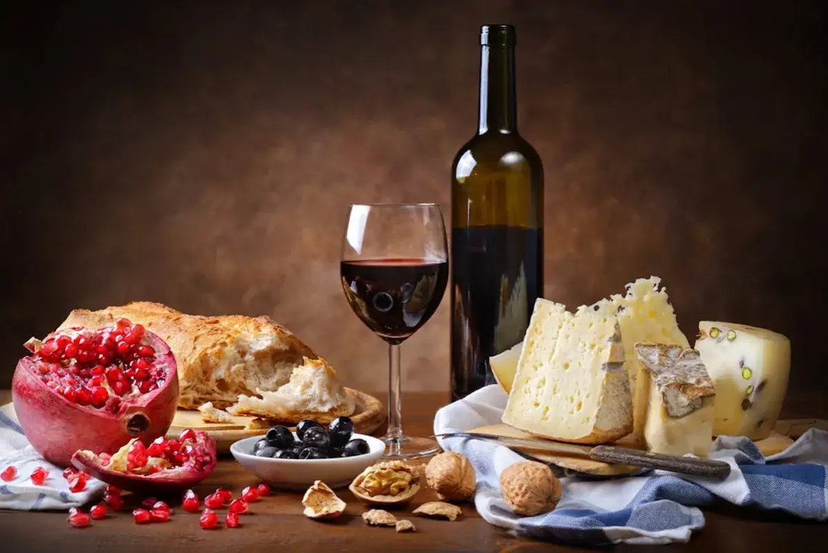 Ein Glas Rotwein, Brot, Granatapfel, Oliven und Käse auf einer rustikalen Tafel. Ein Festmahl für Genießer.