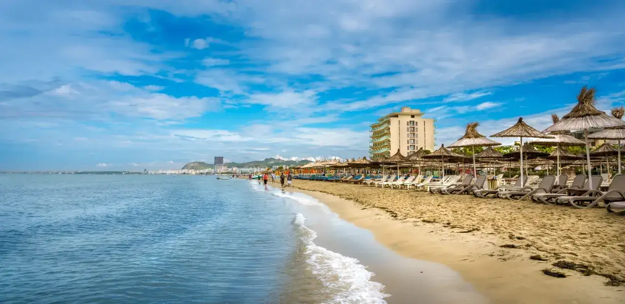 Słoneczna plaża w Durrës, z rzędem leżaków i parasoli, zaprasza do relaksu. W oddali widać miasto i wzgórza, a na pierwszym planie łagodne fale Adriatyku. To idealne miejsce na wakacyjne atrakcje.