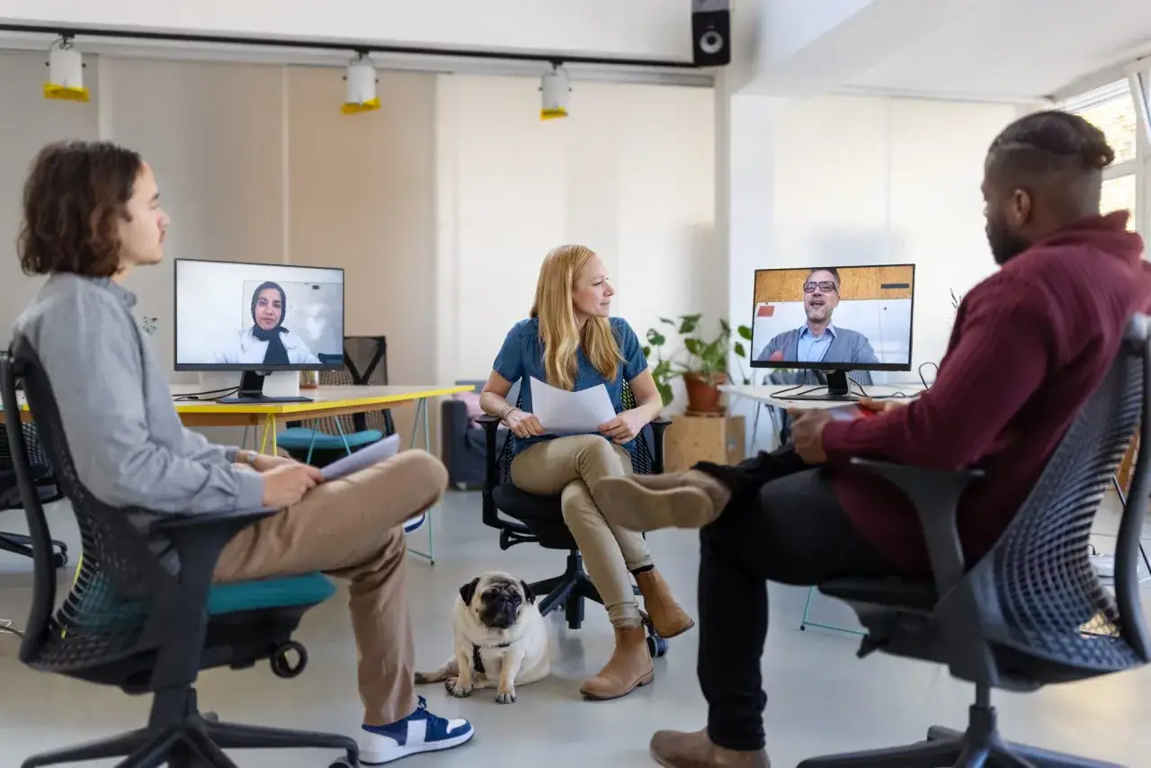 Teammedlemmar jobbar hybrid i ett modernt kontor, med en videokonferens på skärmarna och en söt mops som sällskap.